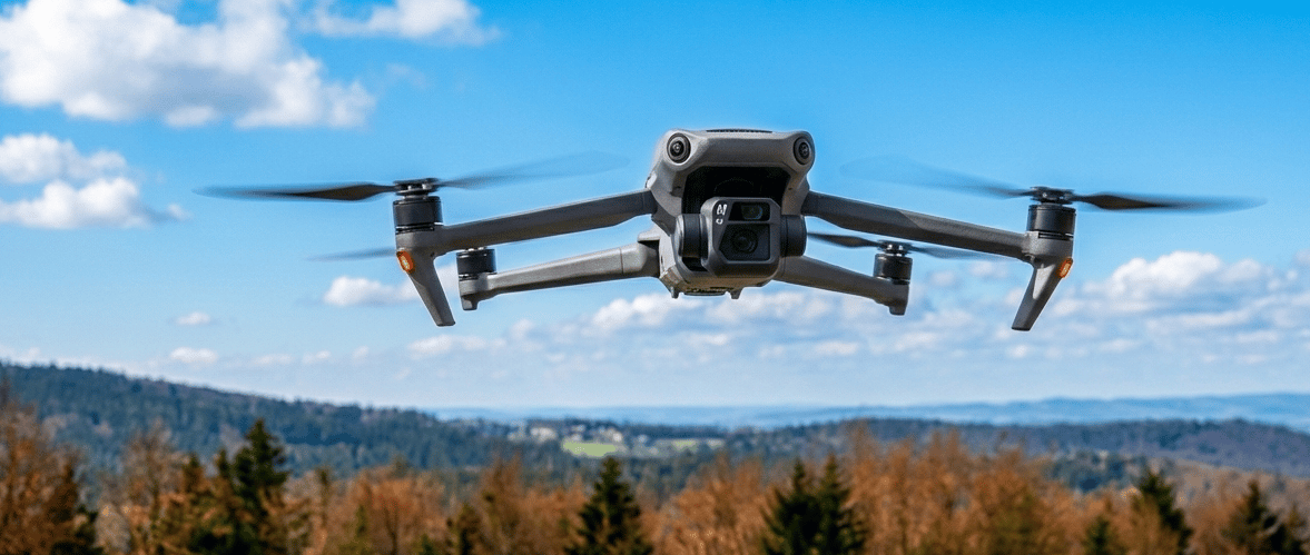 A gray DJI Mavic 3 quadcopter drone hovering in a blue sky over a scenic forest landscape.