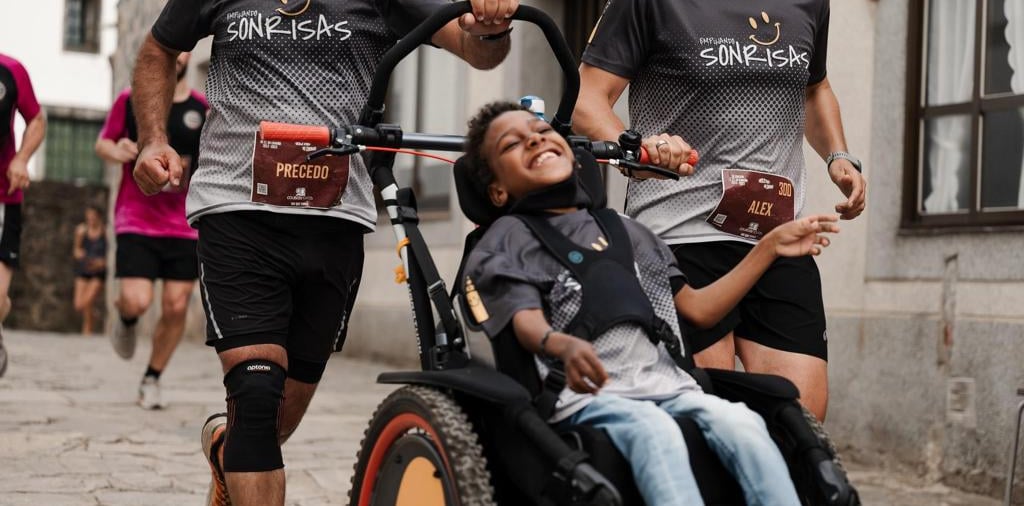 Dos hombres empujan corriendo una silla de ruedas adaptada donde se puede ver a un niño sonriendo