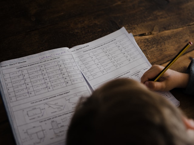 a child's hand holding a pencil and writing on a Maths work book