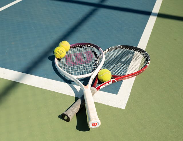 Two tennis rackets and yellow tennis balls on a blue and green outdoor hard court.