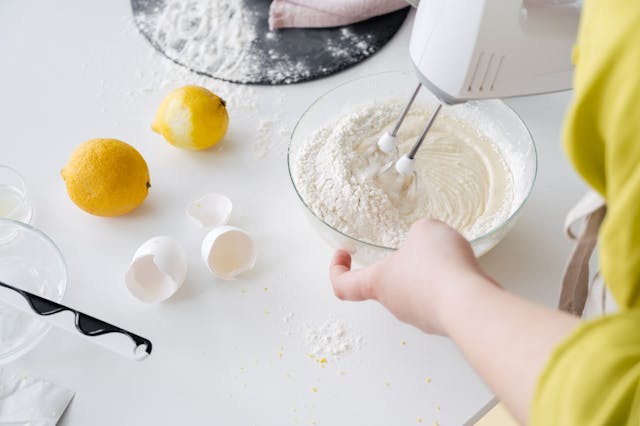 A person using an electric hand mixer to blend cake batter in a glass bowl with fresh lemons and eggshells.