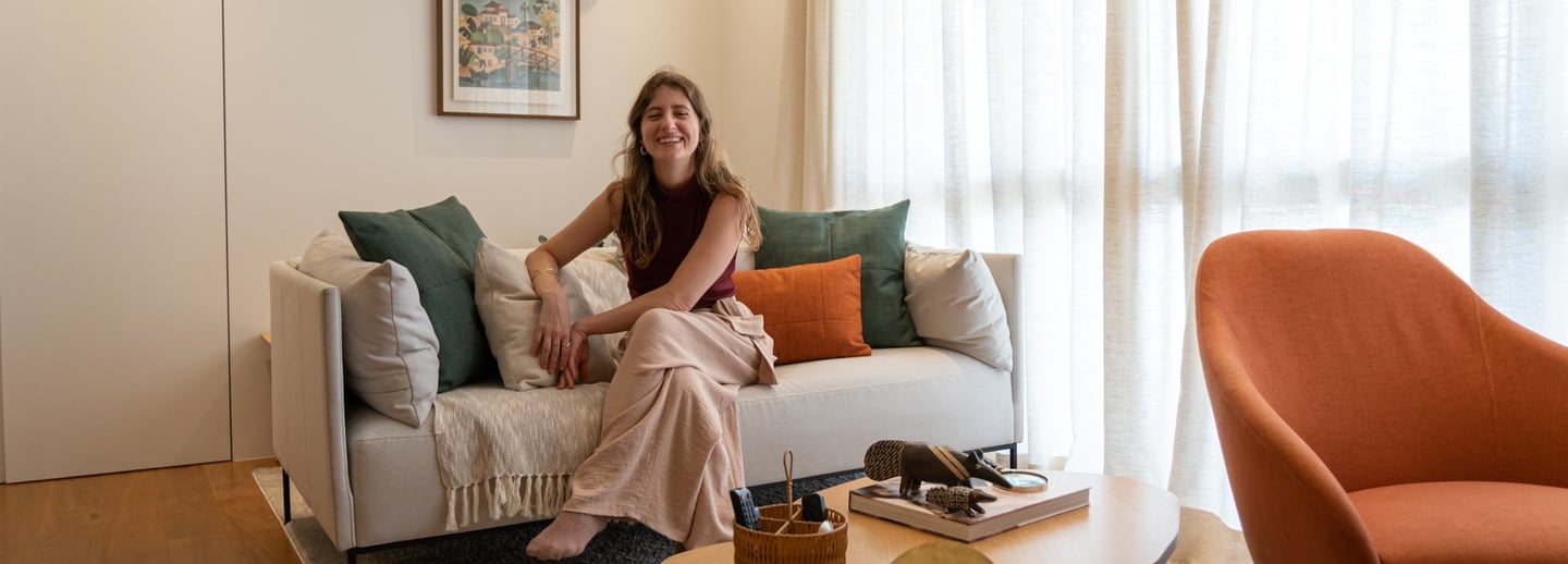 Mulher jovem sorrindo, sentada descalça em um sofá com almofadas coloridas, em uma sala aconchegante