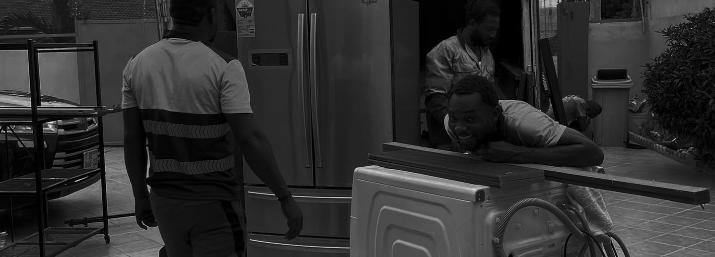 Black and white photograph of three movers efficiently loading large appliances into the open rear o