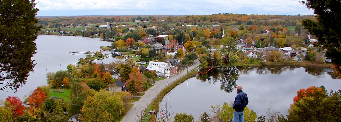 A man looks out from a rocky overlook at a scenic waterfront town with autumn foliage.
