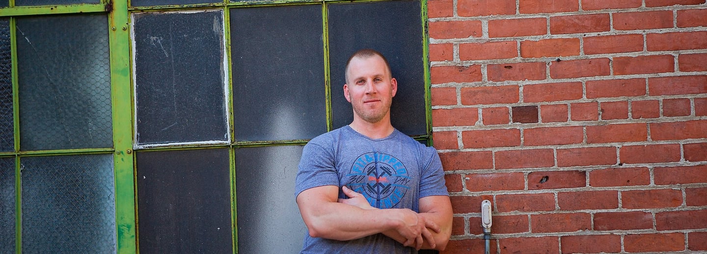 a man posing outside with his arm crossed next to a green window and brick wall