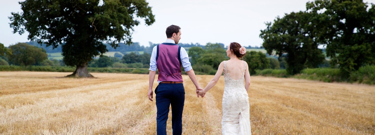 A bride and groom holding hands while walking through a golden rural field after their wedding ceremony.