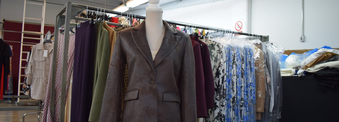 Maniquí con chaqueta corta de polipiel marrón. Fondo con perchas con ropa