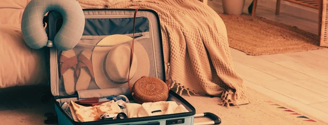 Open suitcase packed with travel essentials like a sun hat, neck pillow, and clothes on a cozy rug.