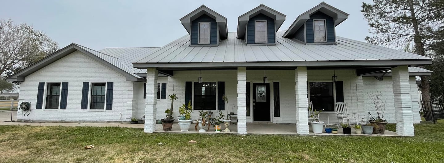 White brick ranch style home with a grey metal roof and three dormer windows on a grassy lawn.
