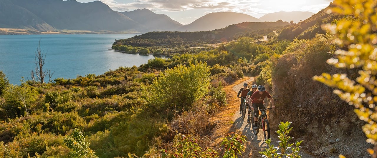 Mountain bikers riding on a scenic dirt trail overlooking New Zealand mountains at sunset.