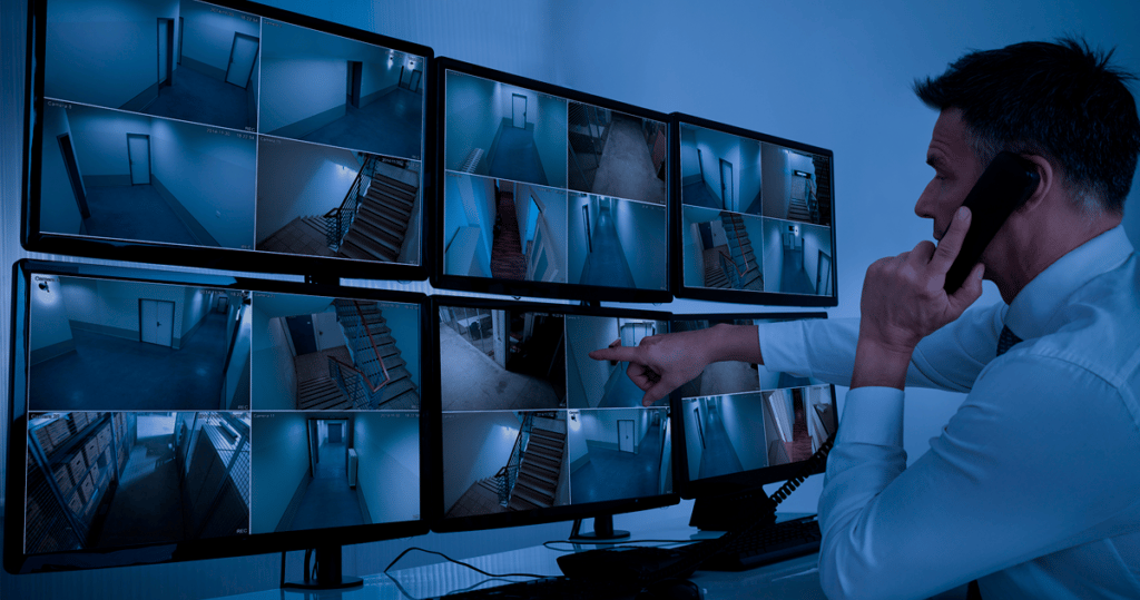 Security guard monitoring multiple CCTV camera feeds on a wall of video surveillance displays.