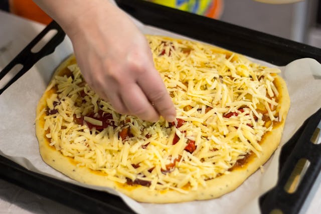 Hand sprinkling shredded cheese on a homemade pizza with tomatoes on a baking sheet.