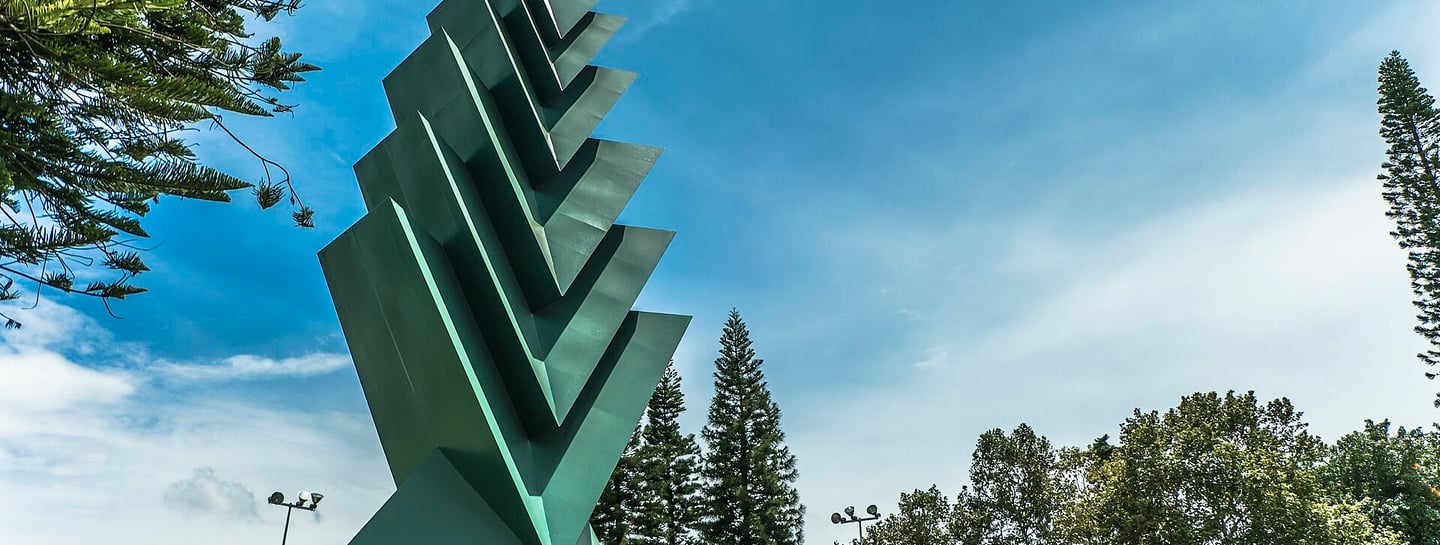 Modern green geometric sculpture stands tall in a public park under a bright blue sky.