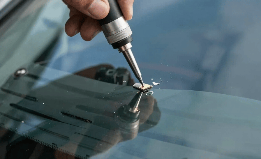 Professional photography of a technician's hand using a precision resin tool to repair a small chip in a car windshield, sharp focus on the glass, reflecting a clear blue North American sky and distant mountain peaks.