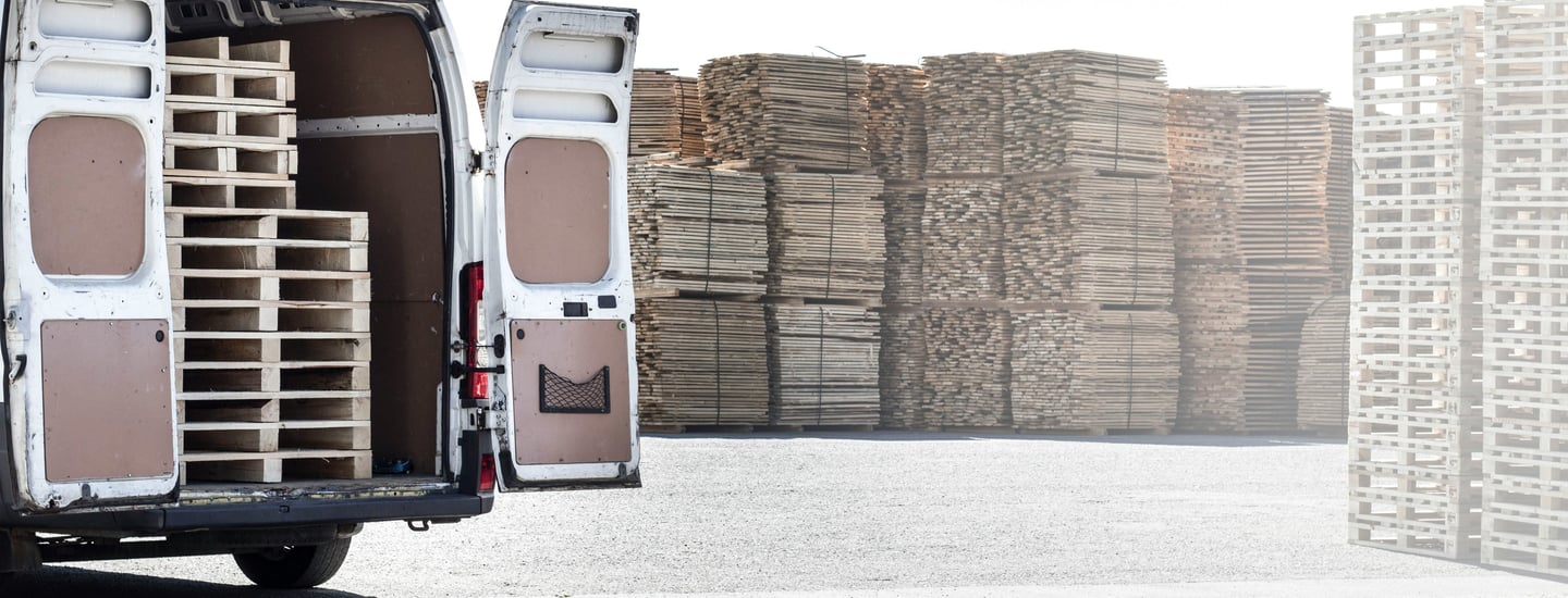 Open van loaded with wooden pallets in an industrial yard, surrounded by stacks of pallets in the background. Represents reli