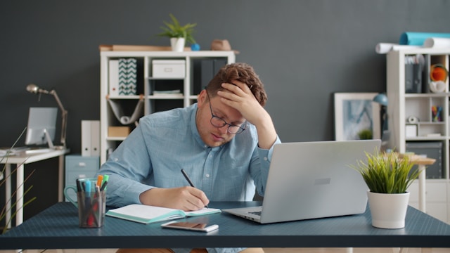 a man sitting at a desk with a laptop and a notebook, thinking how he can save money as a student