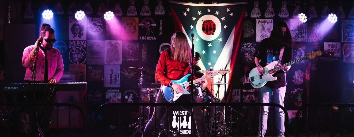 A live rock band performs on a stage with neon lighting and a poster-filled backdrop.