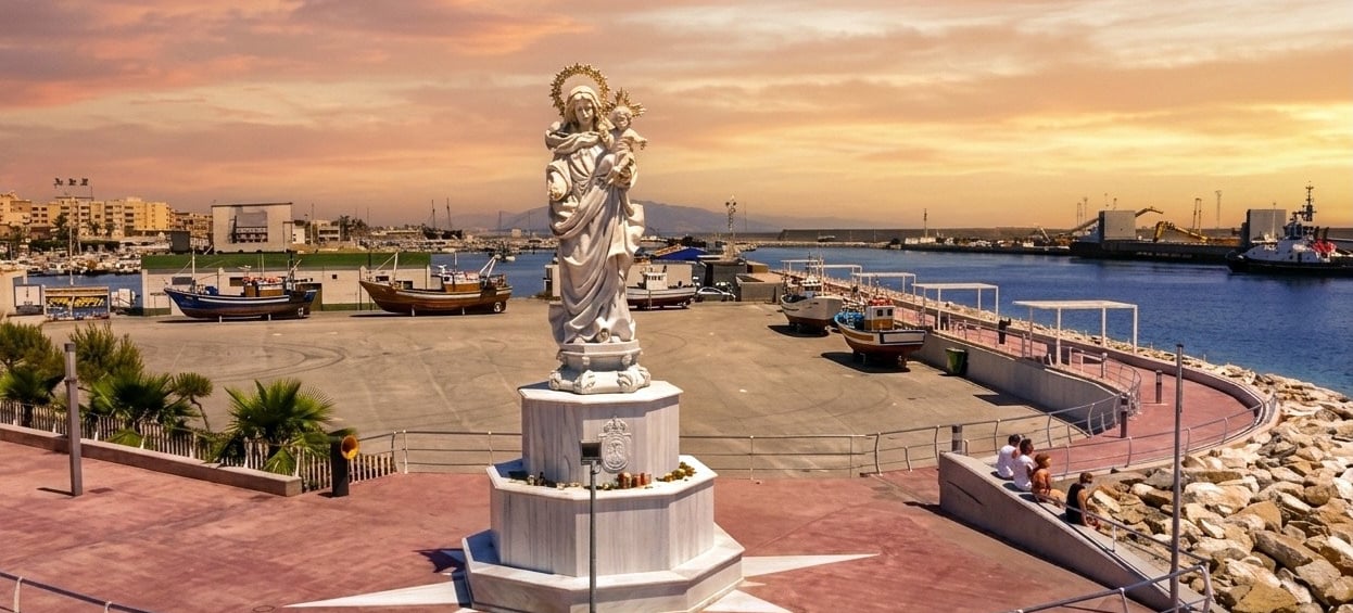 A white marble statue of the Virgin Mary overlooking a coastal fishing harbor at sunset.