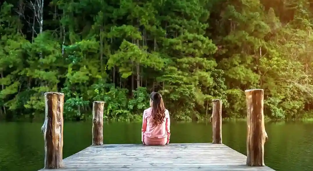 Femme de dos cheveux longs habits roses assise sur un ponton au bord d'une rivière bordée d'arbres