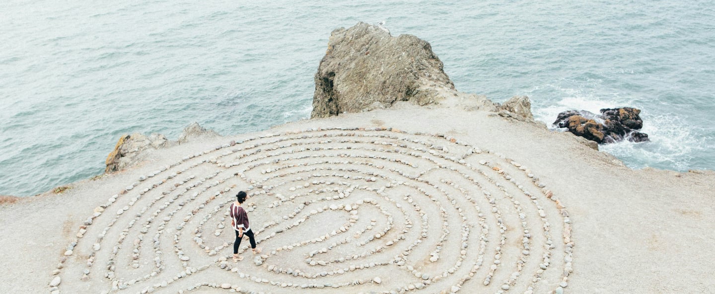 Eine Person ist in der Mitte eines runden Labyrinths an einer Klippe zum offenen Mehr hin.