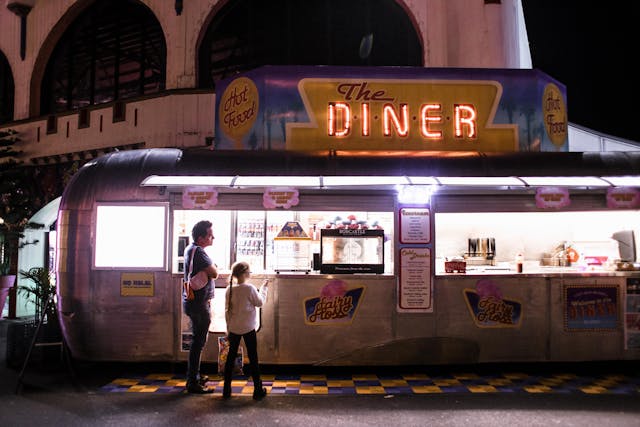 Customers ordering food from a vintage silver Airstream food truck diner with neon signs at night.