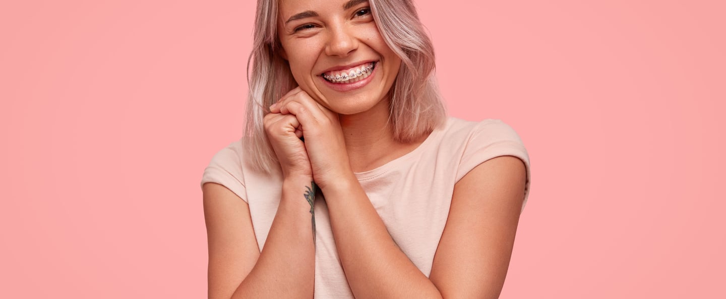 Smiling young woman with silver hair showing dental braces against a pink background.