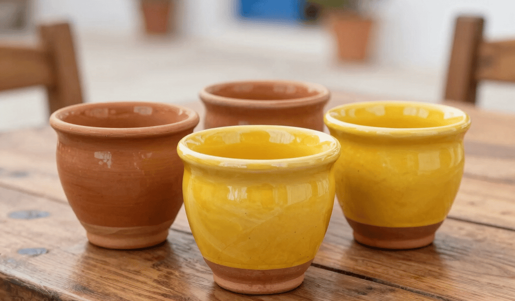 A close-up photograph of vibrant glazed terracotta and honey yellow stoneware ceramic cups placed on a wooden rustic dining table in a Mediterranean courtyard in Turkey.