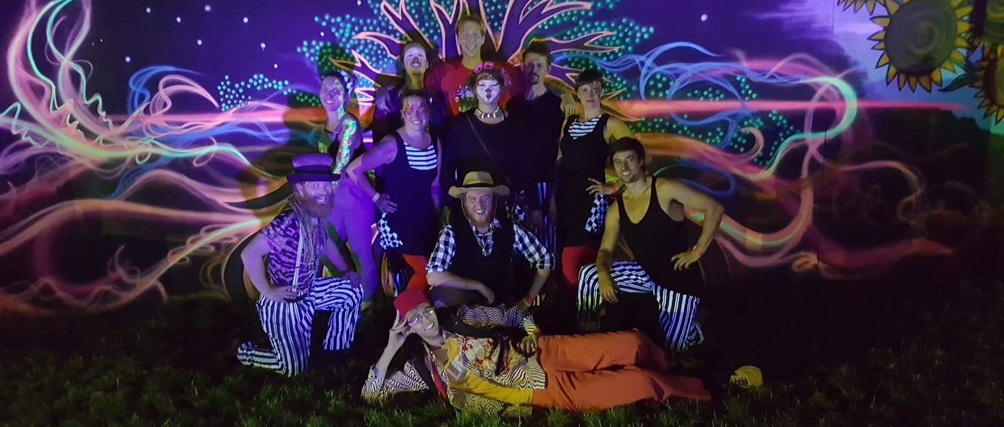 A troupe of circus performers in colourful costumes posing before a vibrant neon projection mural.