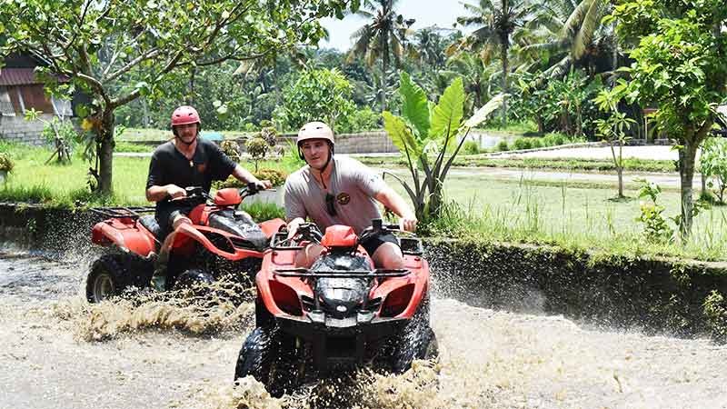 Two men riding red ATVs through a muddy river on a jungle quad bike tour in Bali.