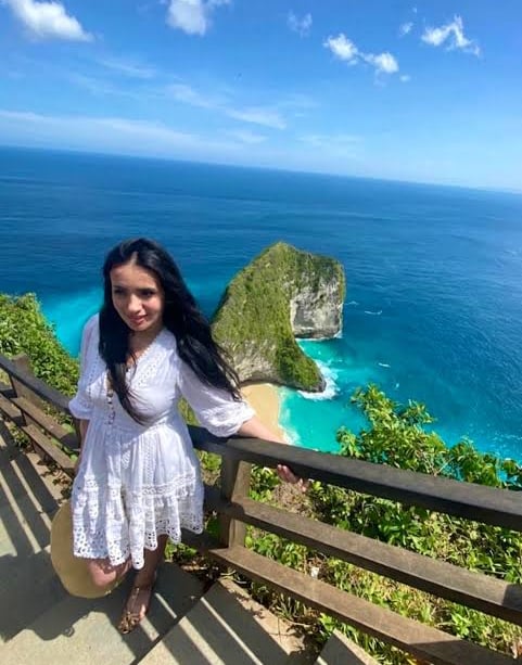 A young woman in a white lace dress poses at Kelingking Beach viewpoint in Nusa Penida, Bali.