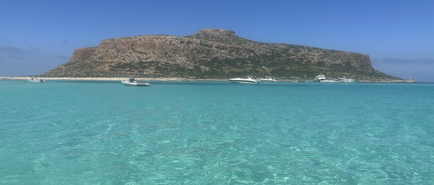 Crystal clear turquoise water at Balos Lagoon in Crete with boats near a rocky island.