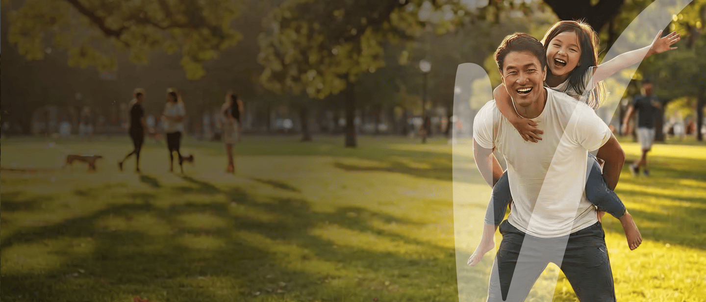 A happy father gives his young daughter a piggyback ride in a sunny, green park.