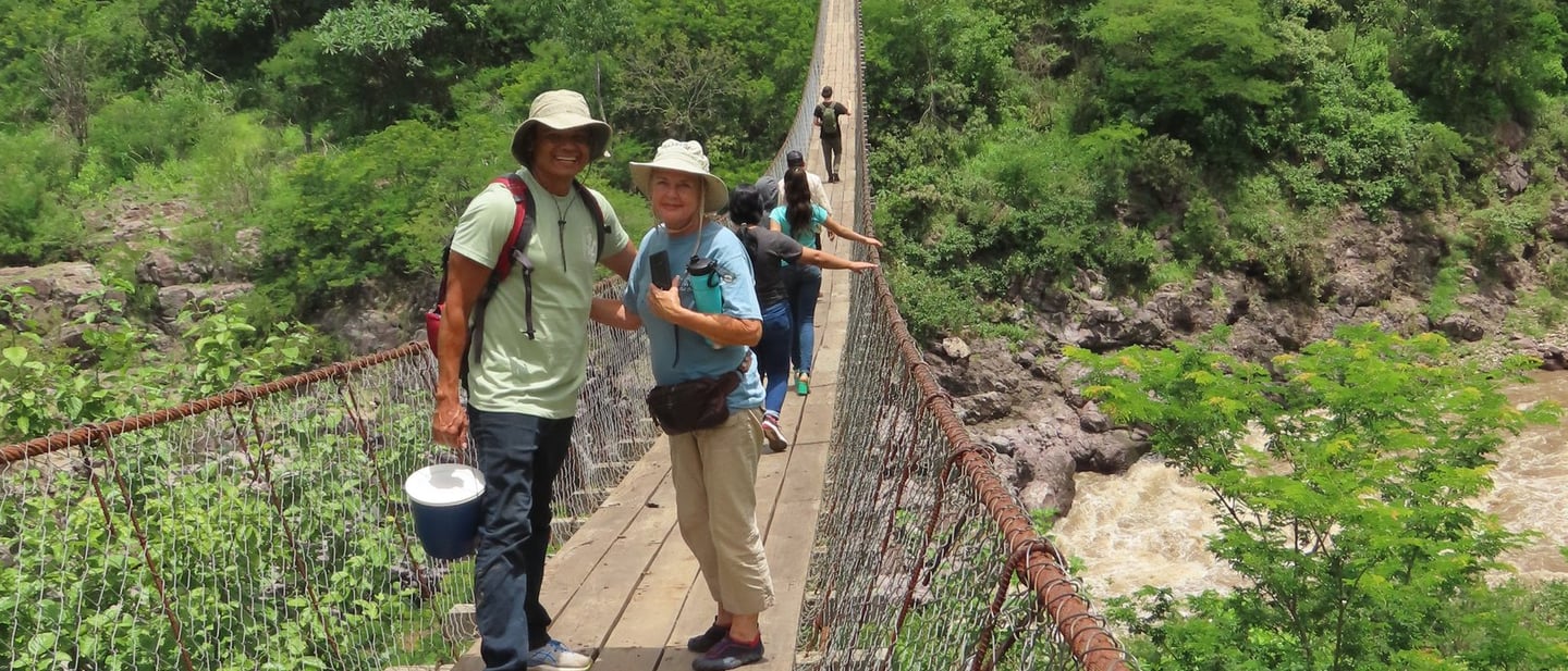 Roger Orozco, and a volunteer smile together as others cross a bridge over rocky terrain below.