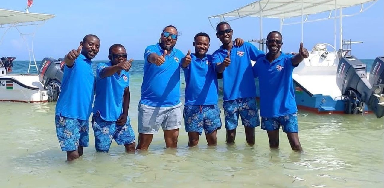 A group of smiling scuba intructors in blue shirts giving thumbs up in the ocean near boats.