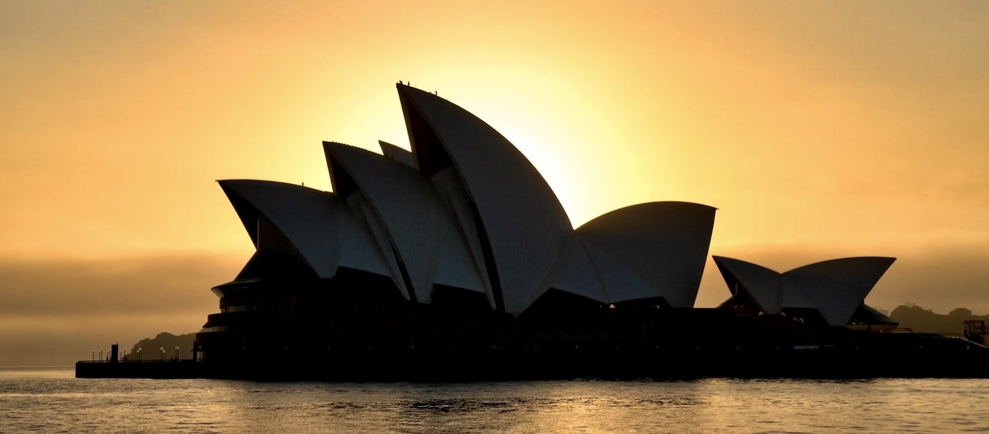 Silhouette of the Sydney Opera House at sunrise against a glowing orange sky over the water.