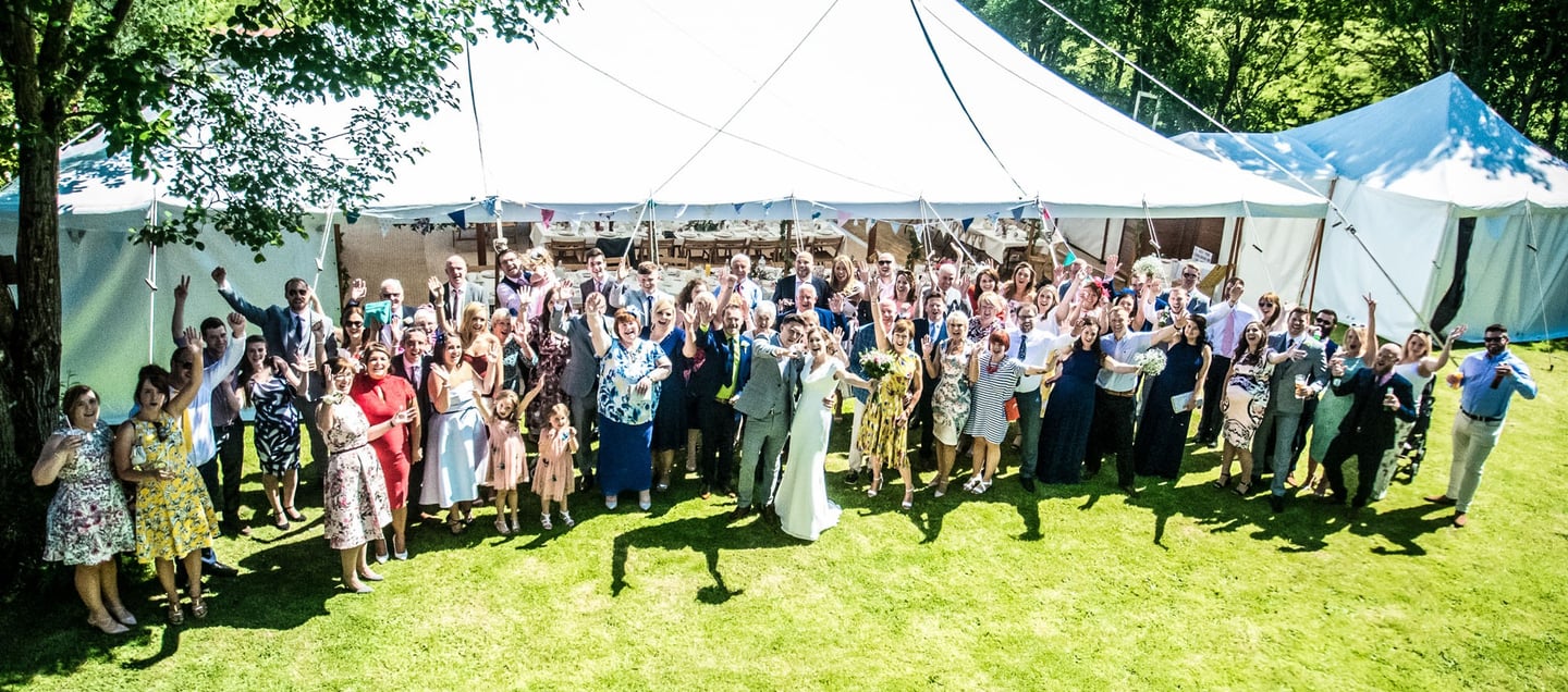 Wedding guests cheering with the bride and groom outside a large white marquee tent on a sunny lawn.