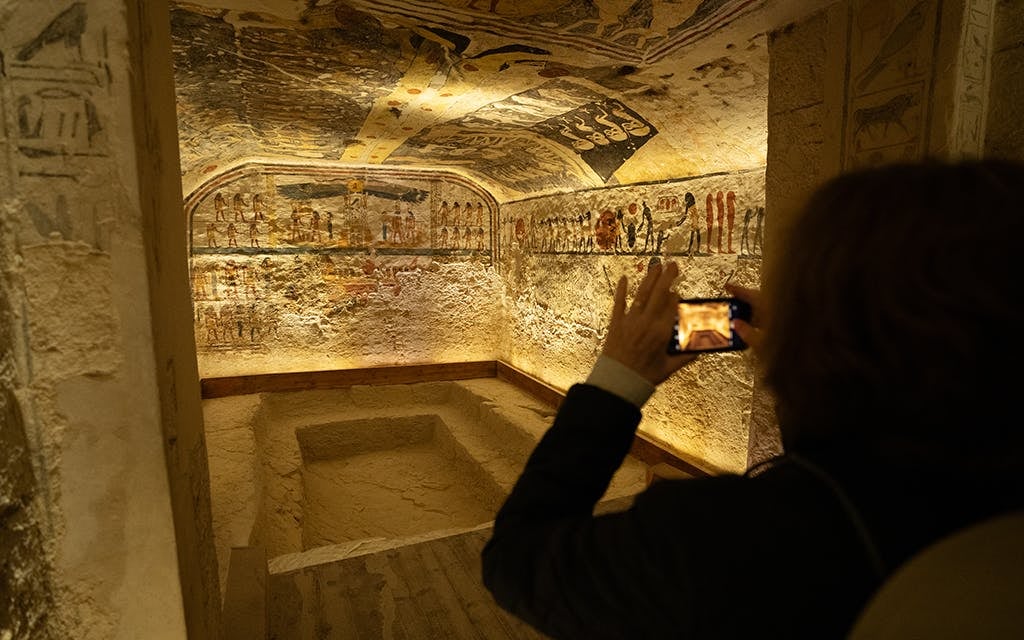 Tourist taking a photo of ancient Egyptian hieroglyphics and wall paintings inside a stone tomb.