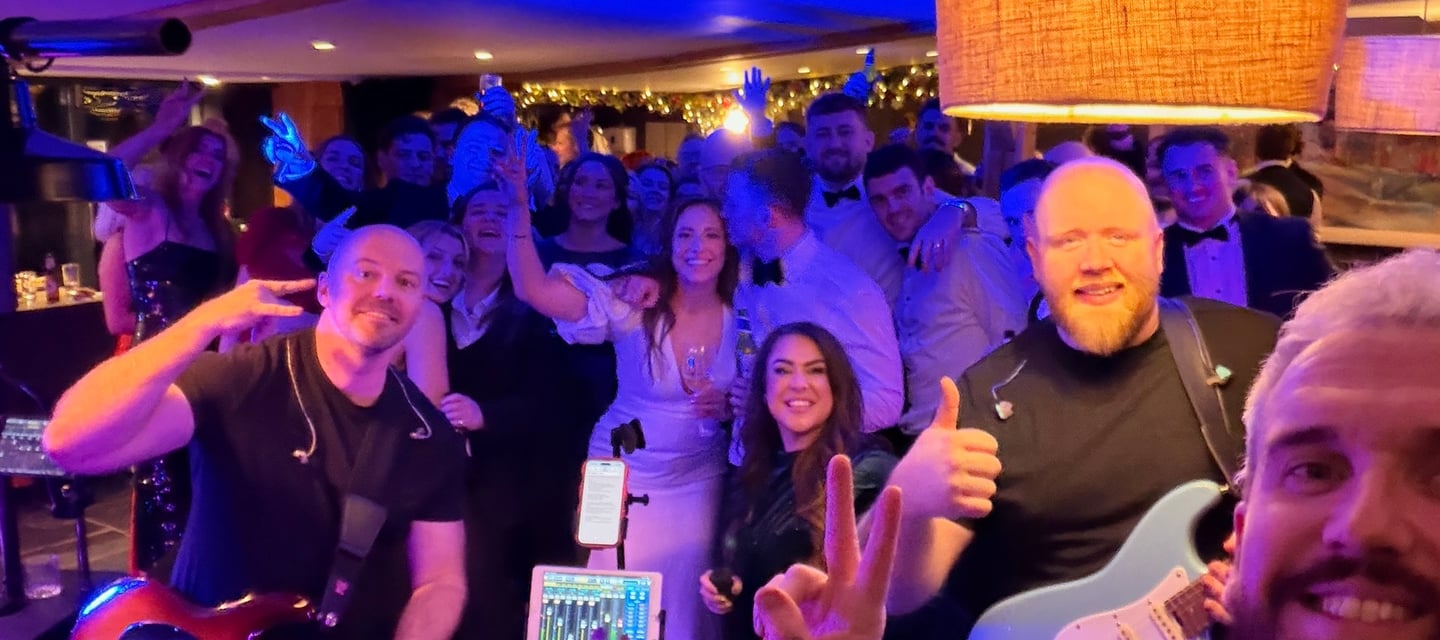 A live wedding band with guitars posing for a selfie with a cheering crowd on a blue lit dance floor.