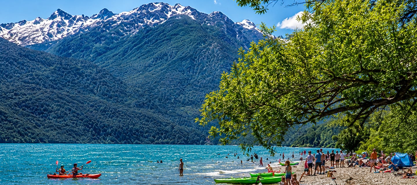 Tourists kayaking and swimming at a sunny mountain lake beach with snow-capped peaks in the background.