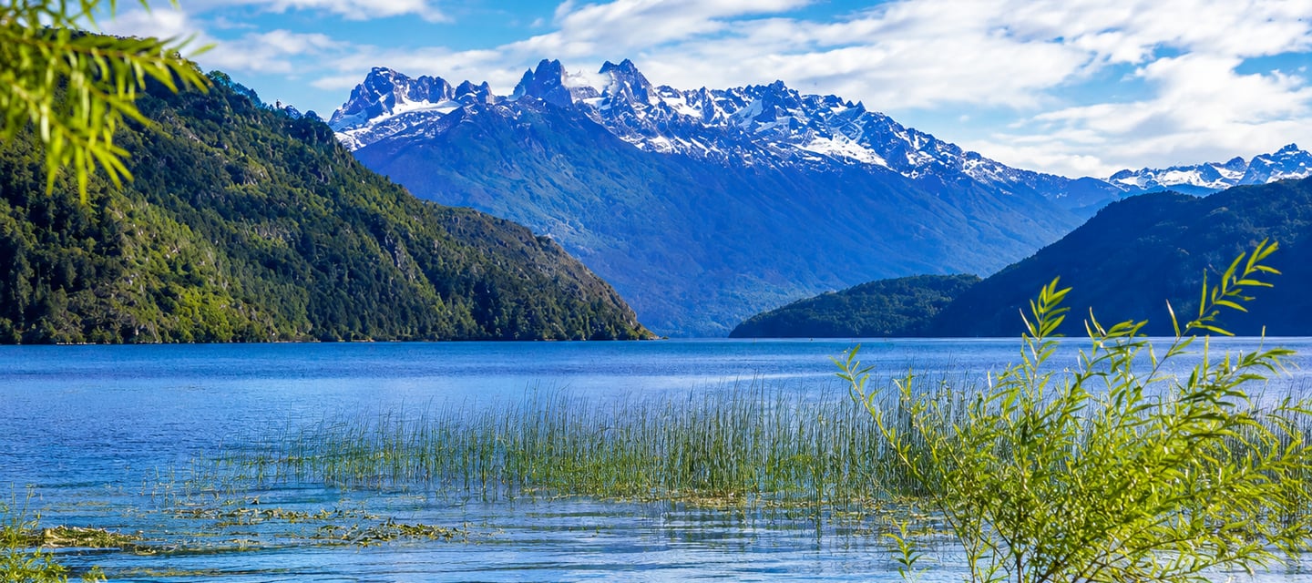 Snow-capped mountain range reflecting in a clear blue lake surrounded by lush green forests.