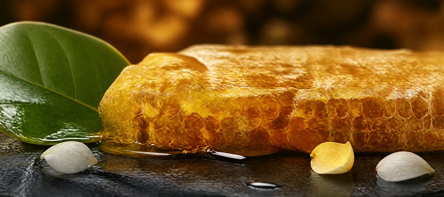 Golden raw honeycomb with dripping honey, green leaf and flower petals on a dark slate surface.