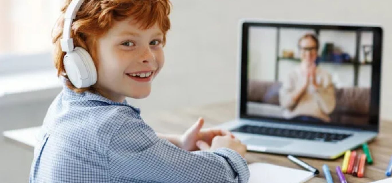 a young boy with headphones and a laptop, on the screen you can see his language tutor, both smiling