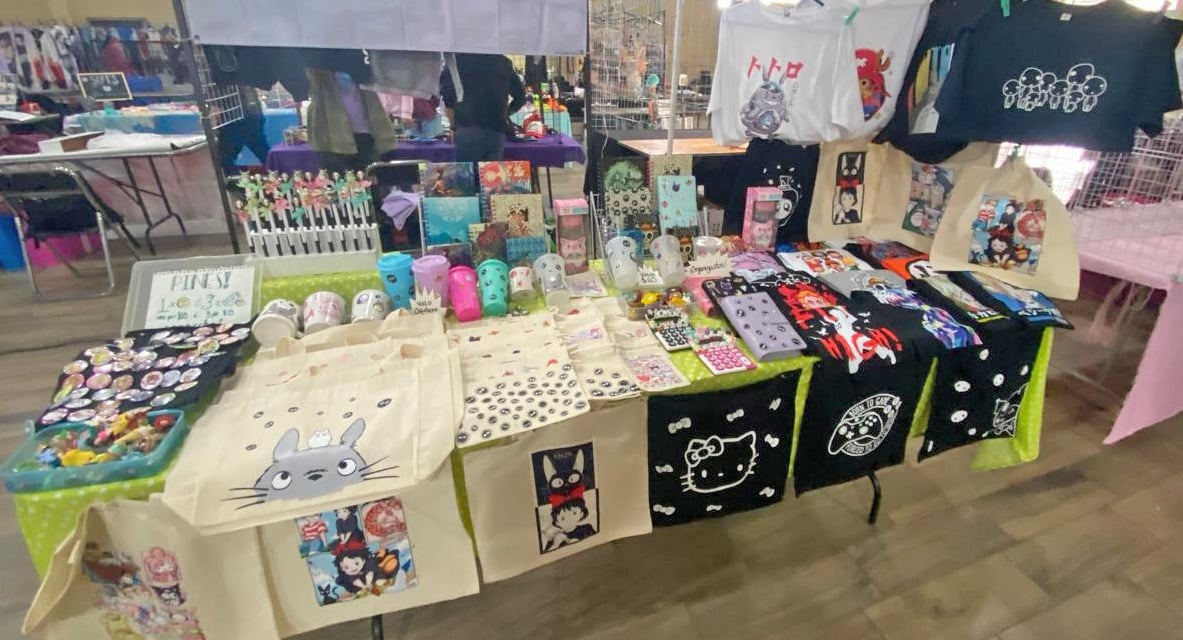 Un puesto de venta de productos de anime con bolsos, playeras,  pines en un bazar