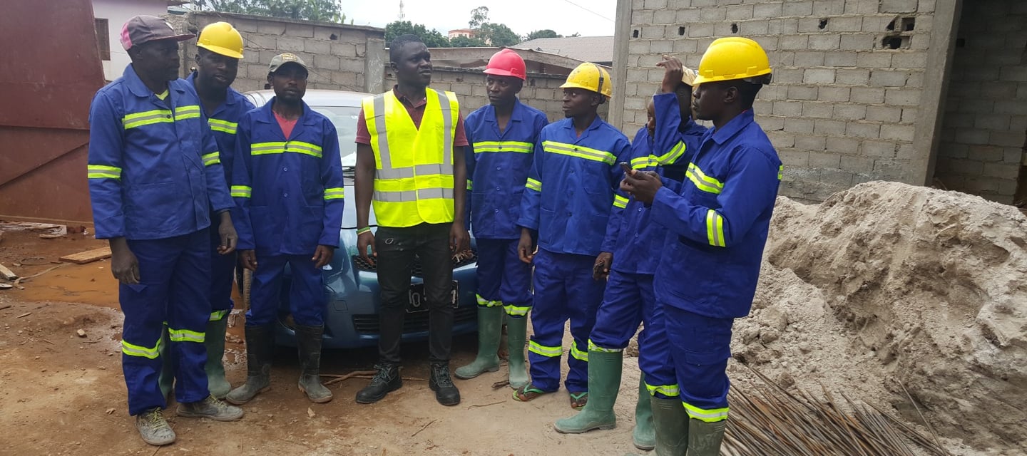 A group of African construction workers wearing blue overalls and hard hats at a building site.