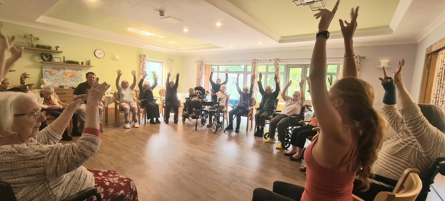 Group of elderly residents taking part in a seated exercise and chair yoga class, led by Lucy