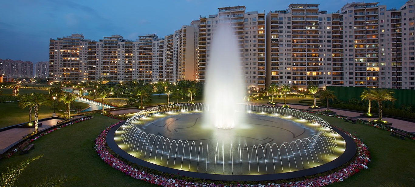 Illuminated circular water fountain in front of luxury high-rise apartment buildings at dusk.
