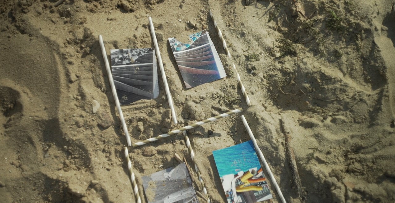 Film still from Excitement by Tina Bikic showing photographs arranged in sand and framed a storyboard with stripped sticks.
