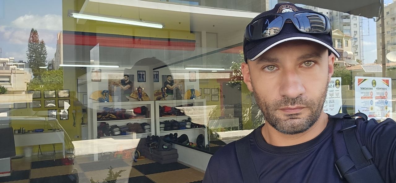 A man in a baseball cap and sunglasses takes a selfie in front of a martial arts studio window.