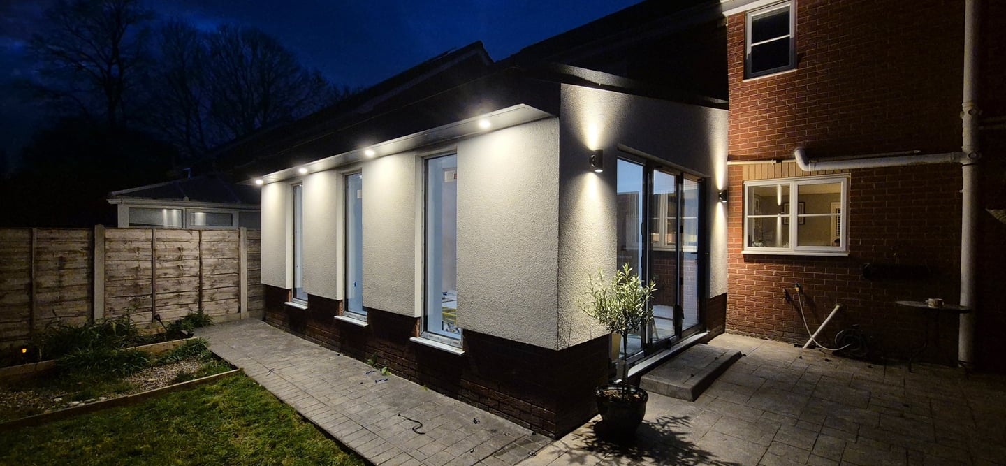 Modern home extension at night with bright soffit spotlights and outdoor wall lighting on white render.