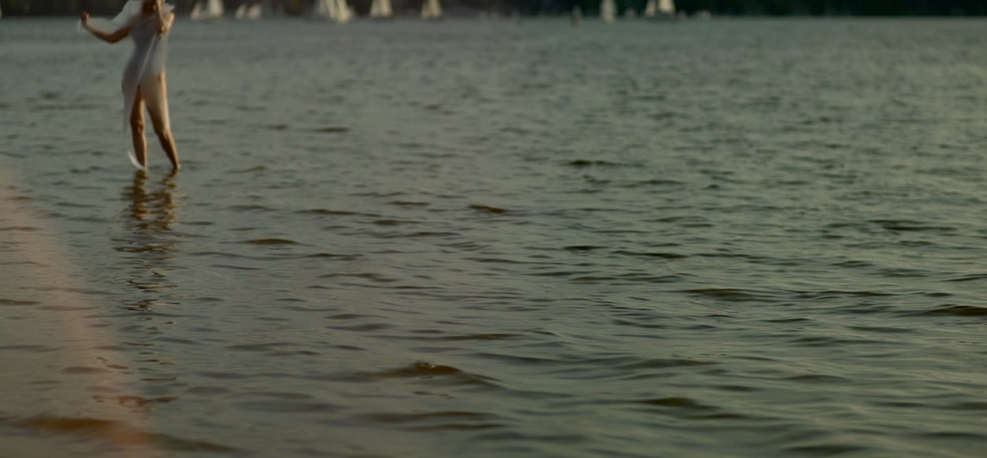 Film still from "Desire for Belonging" by Tina Bikic of a woman dancing through water with sailboats in the distance.